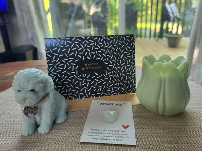 Small dog figurine, birthday card, pocket hug, and green candle on a textured surface.