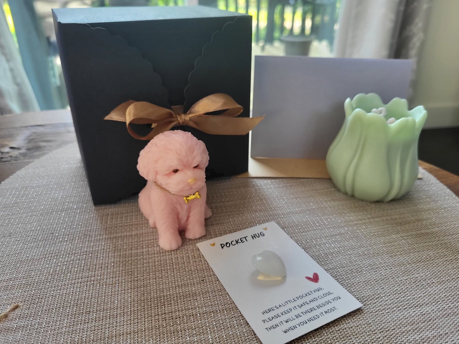 Small pink dog figurine with a gold tag on a textured surface next to a black box and green candle.